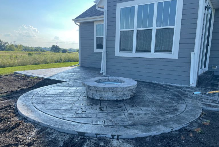 Concrete patio with circular fire pit, featuring stamped design, adjacent to a house, showcasing expert installation by Morici Bros Concrete LLC.