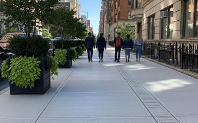 Newly installed concrete sidewalk in an urban setting with pedestrians walking, surrounded by planters and buildings, illustrating durable walkway solutions for property enhancement.